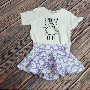 Pode incluir: Uma camiseta branca com um gr&aacute;fico de fantasma e o texto "Spooky Cute" nela. A camiseta &eacute; combinada com uma saia roxa com um padr&atilde;o de fantasma.