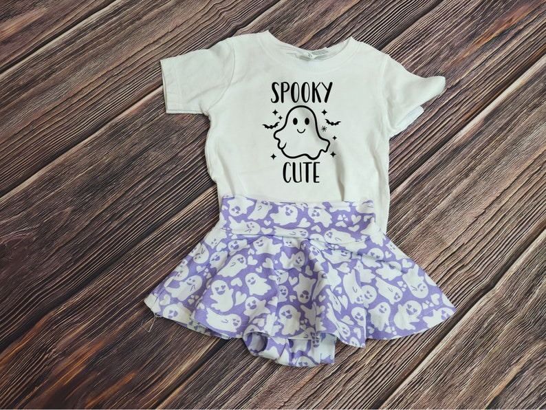 Pode incluir: Uma camiseta branca com um gr&aacute;fico de fantasma e o texto "Spooky Cute" nela. A camiseta &eacute; combinada com uma saia roxa com um padr&atilde;o de fantasma.