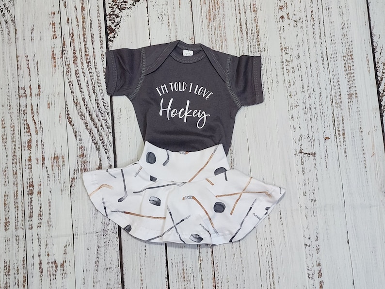 May include: A gray baby bodysuit and skirt set. The bodysuit has the text "I'M TOLD I LOVE Hockey" in white script. The skirt is white with a pattern of hockey sticks and pucks in brown and gray.