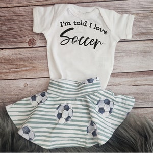 May include: White baby onesie with the text "I'm told I love Soccer" in black. A green and white striped skirt with a soccer ball pattern.