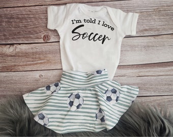 May include: White baby onesie with the text "I'm told I love Soccer" in black. A green and white striped skirt with a soccer ball pattern.