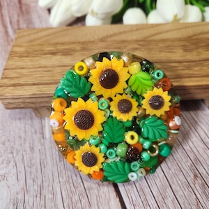May include: A round, resin-based sunflower brooch. The brooch features several yellow sunflowers with brown centers, green leaves, and colorful beads. The brooch is set on a wooden surface.