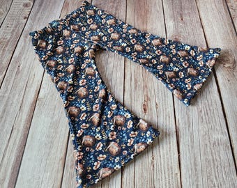May include: Navy blue bell-bottom pants with a repeating pattern of footballs and small floral elements in shades of brown, peach, and white. The pants are made of a soft, stretchy fabric, perfect for comfort and style.