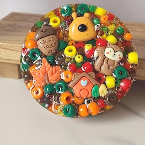 May include: A round, colorful resin art piece with autumn-themed embellishments. It features a bear, squirrel, acorn, leaf, and house, surrounded by beads in orange, green, yellow, and red. The piece is set on a wooden surface.