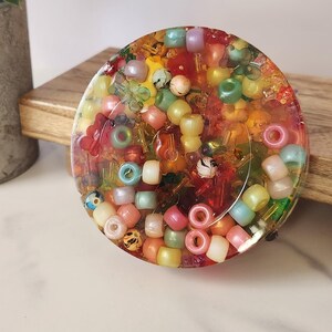 K&ouml;nnte beinhalten: Ein runder, farbenfroher Untersetzer aus Kunstharz, gef&uuml;llt mit einer Auswahl an Perlen in verschiedenen Formen und Gr&ouml;&szlig;en. Die Perlen sind in den Farbt&ouml;nen Rot, Gelb, Gr&uuml;n, Rosa und Blau gehalten. Der Untersetzer hat einen Durchmesser von etwa 10 cm.