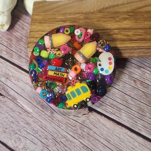 May include: A round resin art piece with a school theme. It features yellow pencils, a yellow school bus, and a palette. The words "ART MATH READING" are visible on a small book. Colorful beads and small embellishments fill the piece.