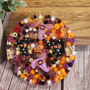 May include: A round, resin coaster with a Halloween theme. The coaster features orange, purple, and white beads, with small witch hats and black and white star-covered figures. The coaster is set on a wooden surface.
