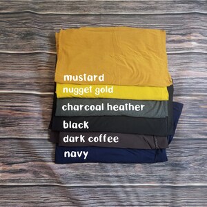 May include: A stack of six t-shirts in different colors, from top to bottom: mustard, nugget gold, charcoal heather, black, dark coffee, and navy.