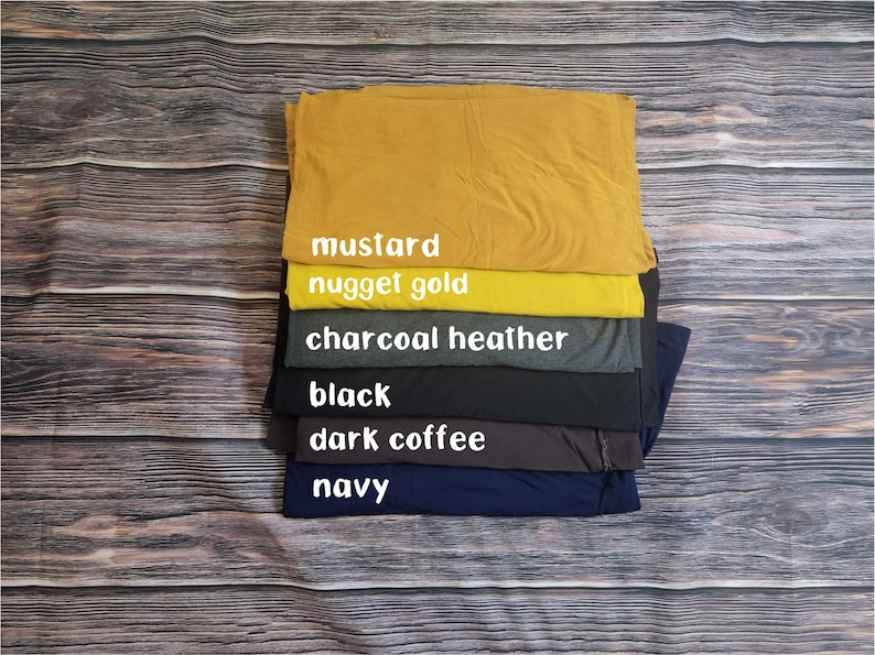 May include: A stack of six t-shirts in different colors, from top to bottom: mustard, nugget gold, charcoal heather, black, dark coffee, and navy.