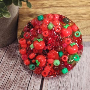 May include: A round, resin-based decorative piece with a vibrant mix of red beads, small red apple-shaped charms, and green accents. The design includes a central rose and various sizes of red beads. The piece is set on a wooden surface.