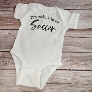 May include: White baby onesie with black text that reads "I'm told I love Soccer".