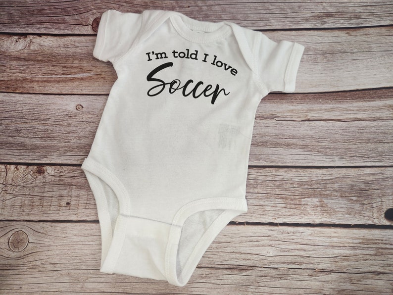 May include: White baby onesie with black text that reads "I'm told I love Soccer".