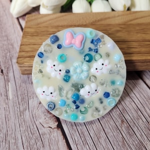 May include: A circular wax melt with a light base, decorated with smiling cloud shapes, a pink bow, and blue and clear embellishments. The melt is displayed on a wooden surface.