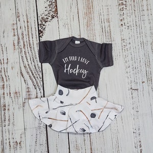 May include: A gray baby bodysuit with the text "I'M TOLD I LOVE Hockey" paired with a white skirt featuring a hockey stick and puck pattern. The outfit is designed for infants and is displayed on a light wood surface.