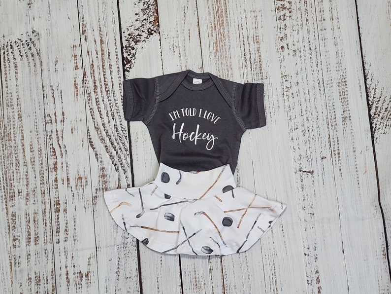 May include: A gray baby bodysuit with the text "I'M TOLD I LOVE Hockey" paired with a white skirt featuring a hockey stick and puck pattern. The outfit is designed for infants and is displayed on a light wood surface.