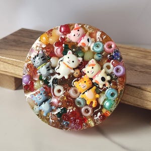 Peut inclure: Une pièce d'art en résine ronde avec plusieurs petites figurines de chats de différentes couleurs, entourées de perles colorées et d'autres éléments décoratifs. Posée sur une surface en bois.