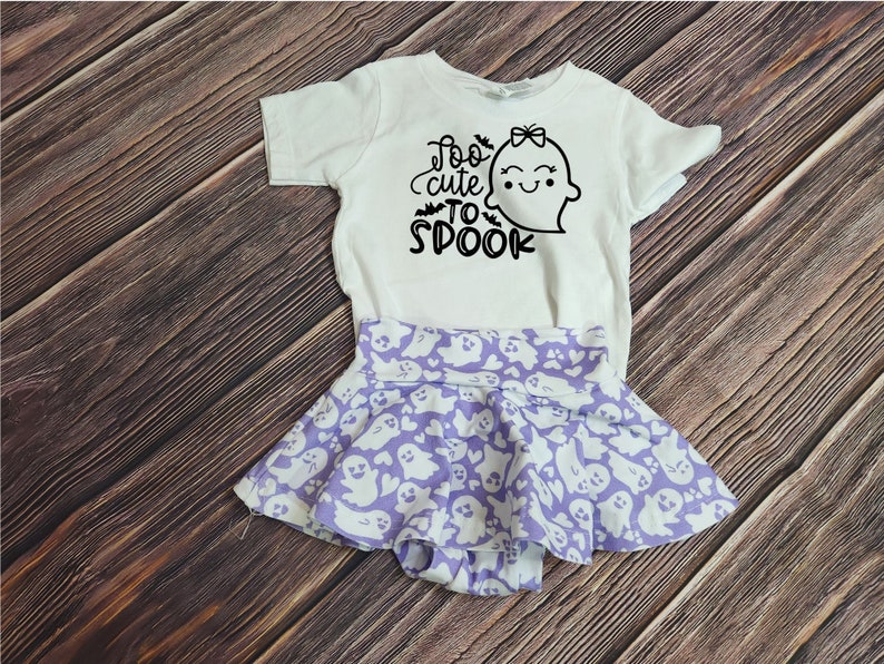 May include: A white baby onesie with a cute ghost graphic and the text "Too cute to spook". The onesie is paired with a purple skirt with a white ghost pattern.