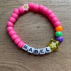 May include: Pink beaded bracelet with the name "MABEL" spelled out in white letter beads. The bracelet also features a yellow star charm, a purple bead, a green bead, an orange bead, and a pink flower bead with a yellow smiley face.