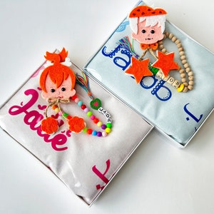 May include: Two personalized baby blankets in gift boxes. One blanket is light blue with the name "Jacob" and a wooden bead necklace with a yellow bead and orange star charms. The other blanket is white with pink lettering and the name "Jade" and a colorful beaded necklace with a green heart charm.