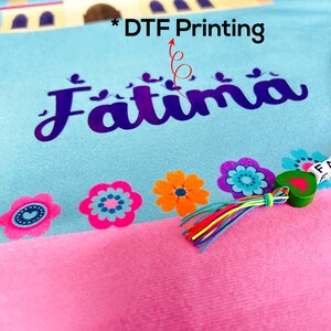 Puede incluir: Una alfombra de oraci&oacute;n azul claro y rosa con un dise&ntilde;o de mezquita y el nombre "Fatima" impreso en ella. La alfombra tiene un borla colorido y un peque&ntilde;o amuleto de coraz&oacute;n verde.