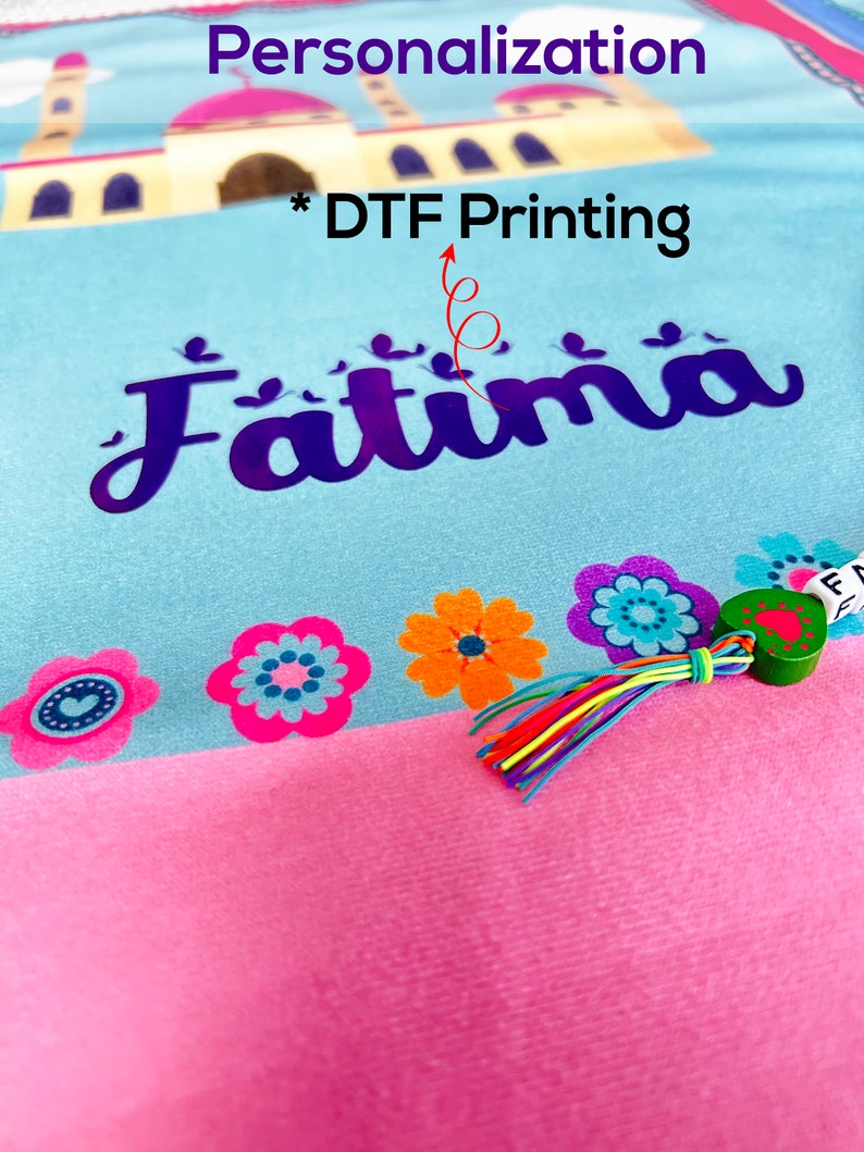 Puede incluir: Una alfombra de oraci&oacute;n azul claro y rosa con un dise&ntilde;o de mezquita y el nombre "Fatima" impreso en ella. La alfombra tiene un borla colorido y un peque&ntilde;o amuleto de coraz&oacute;n verde.