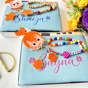 Puede incluir: Dos cajas de regalo azules y blancas con nombres bordados personalizados, "Sumeja" y "Zayna". Cada caja incluye un personaje de fieltro, un collar de cuentas de madera y una pulsera de cuentas de colores.