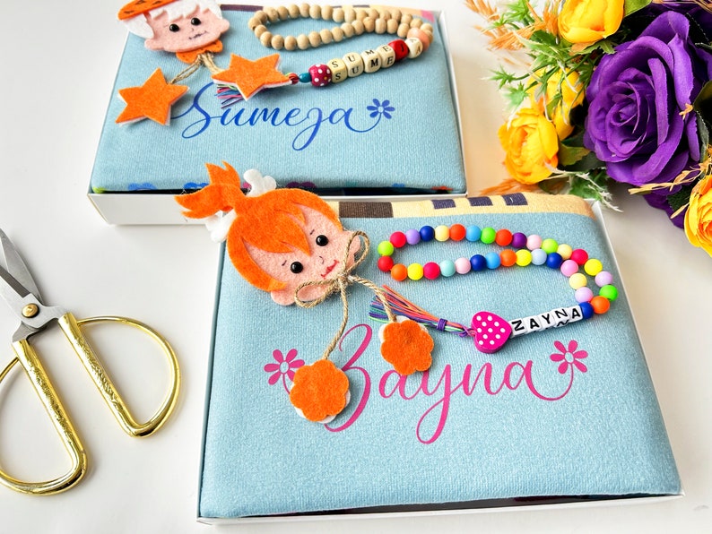 Puede incluir: Dos cajas de regalo azules y blancas con nombres bordados personalizados, "Sumeja" y "Zayna". Cada caja incluye un personaje de fieltro, un collar de cuentas de madera y una pulsera de cuentas de colores.