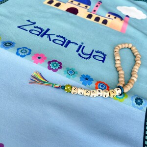 Puede incluir: Una alfombra de oraci&oacute;n azul claro con un dise&ntilde;o de mezquita colorido y el nombre "Zakariya" impreso en azul. Un collar de cuentas de oraci&oacute;n de madera y un amuleto de nombre con cuentas de colores est&aacute;n sobre la alfombra.