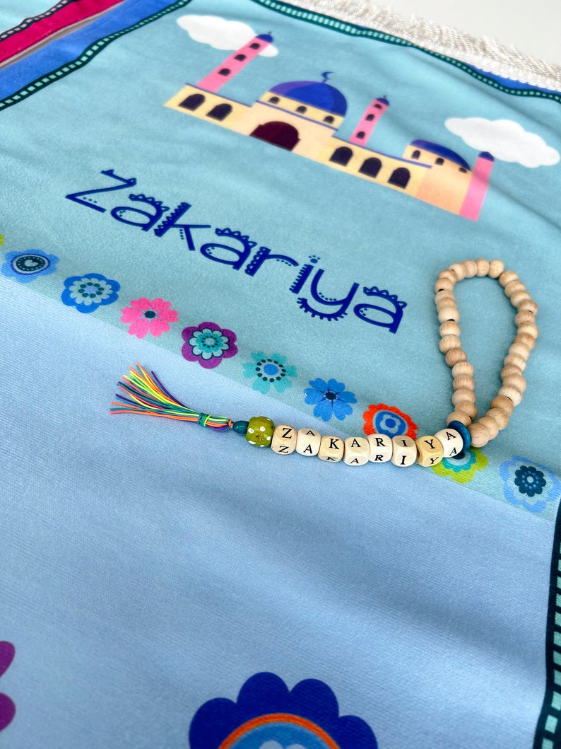 Puede incluir: Una alfombra de oraci&oacute;n azul claro con un dise&ntilde;o de mezquita colorido y el nombre "Zakariya" impreso en azul. Un collar de cuentas de oraci&oacute;n de madera y un amuleto de nombre con cuentas de colores est&aacute;n sobre la alfombra.