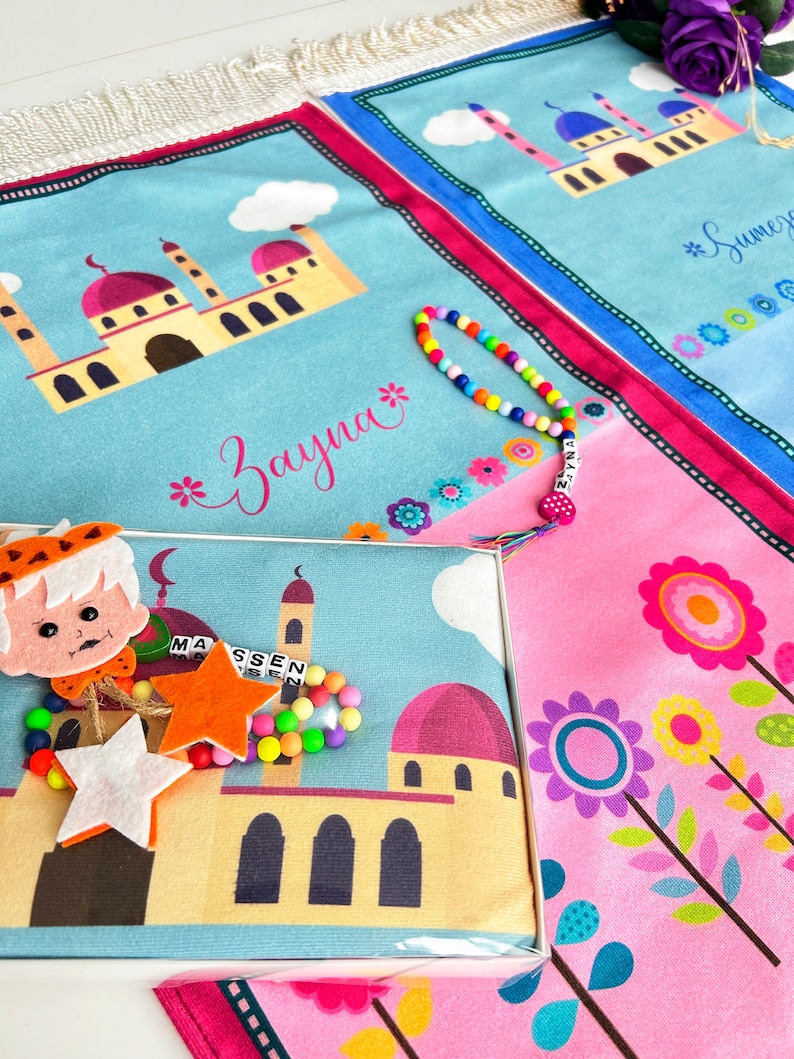 Puede incluir: Dos alfombras de oraci&oacute;n azules y rosas con un dise&ntilde;o de mezquita y los nombres "Zayna" y "Sumey" impresos en ellas. Una estrella blanca y naranja, un brazalete de cuentas de colores y una mu&ntilde;eca de fieltro est&aacute;n encima de la alfombra azul.