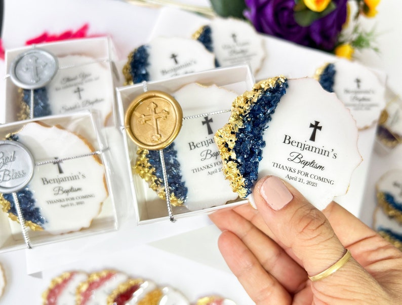 May include: A set of white and gold cookies with blue and gold accents. The cookies are decorated with a cross and the text "Benjamin's Baptism Thanks for Coming April 21, 2023".