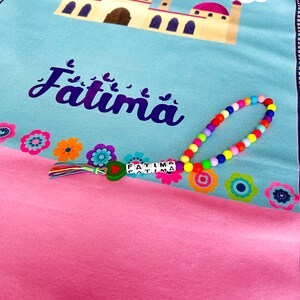 Puede incluir: Una alfombra de oraci&oacute;n rosa y azul con un dise&ntilde;o de mezquita y el nombre "Fatima" impreso en ella. Un collar de cuentas de colores con el nombre "Fatima" deletreado en &eacute;l est&aacute; encima de la alfombra.