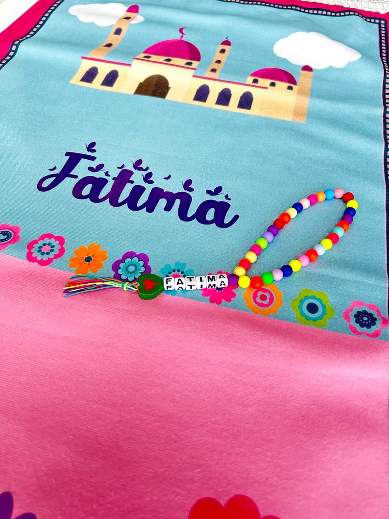 Puede incluir: Una alfombra de oraci&oacute;n rosa y azul con un dise&ntilde;o de mezquita y el nombre "Fatima" impreso en ella. Un collar de cuentas de colores con el nombre "Fatima" deletreado en &eacute;l est&aacute; encima de la alfombra.