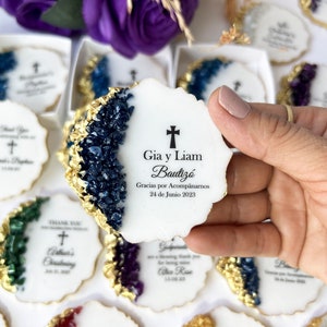 May include: White round cookies with gold foil accents and blue, green, and purple crystal-like designs. The cookies have a cross and text that reads "Gia y Liam Bautiz&oacute; Gracias por Acomp&aacute;narnos 24 de Junio 2023".