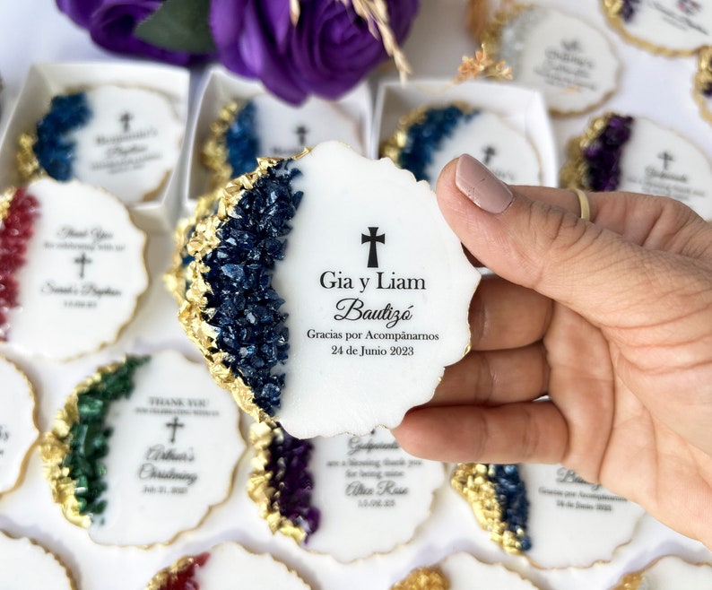 May include: White round cookies with gold foil accents and blue, green, and purple crystal-like designs. The cookies have a cross and text that reads "Gia y Liam Bautiz&oacute; Gracias por Acomp&aacute;narnos 24 de Junio 2023".