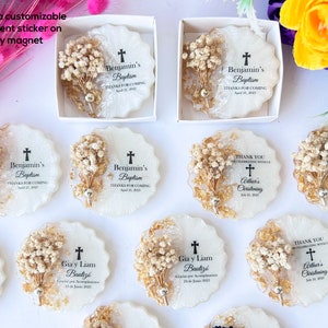 May include: White epoxy magnets with a scalloped edge, decorated with dried flowers and gold accents. Each magnet features a cross and personalized text for a baptism or christening. The magnets are packaged in white boxes.