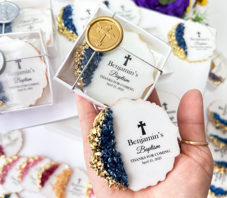 May include: White favor boxes with blue and gold accents, containing cookies decorated with a cross and the text "Benjamin's Baptism Thanks for Coming April 21, 2023".