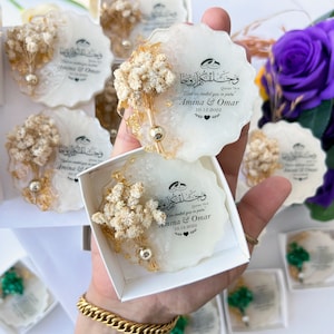 May include: White and gold resin coasters with dried floral accents. Each coaster features calligraphy and the names "Amina & Omar" with a date. The coasters are packaged in small white boxes, ideal for wedding favours.
