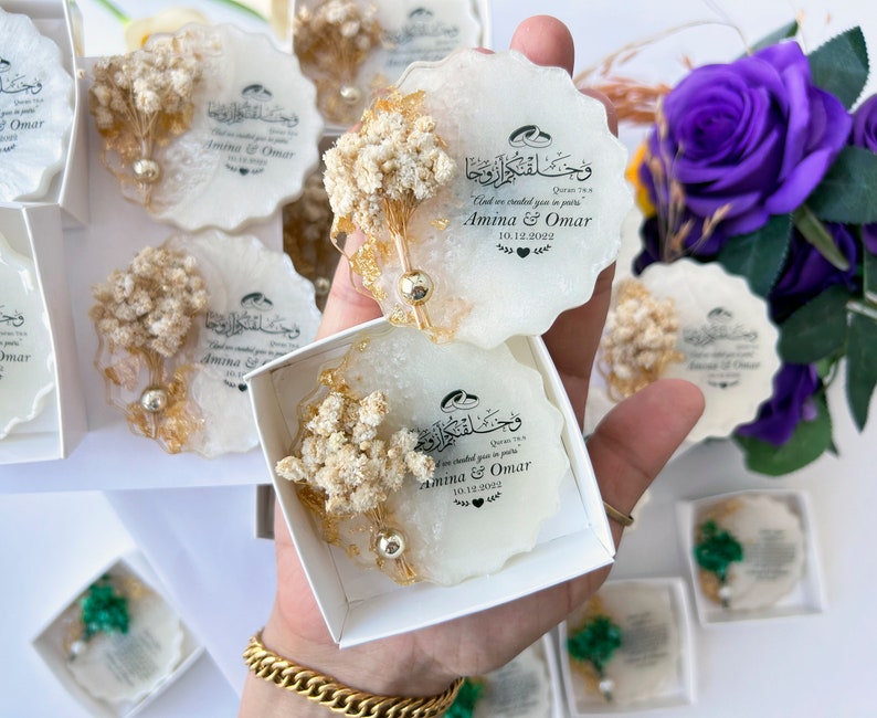 May include: White and gold resin coasters with dried floral accents. Each coaster features calligraphy and the names "Amina & Omar" with a date. The coasters are packaged in small white boxes, ideal for wedding favours.