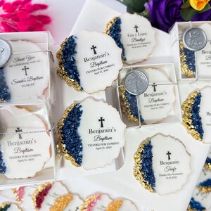 May include: A collection of white cookies decorated with gold and blue frosting, featuring a cross and the text "Benjamin's Baptism" with the date. The cookies are packaged in clear boxes with a silver seal.