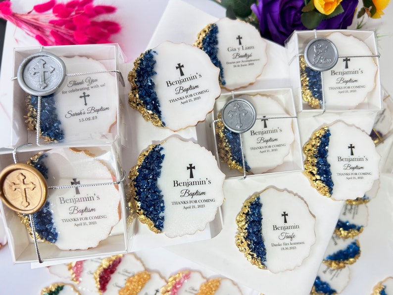 May include: A collection of white cookies decorated with gold and blue frosting, featuring a cross and the text "Benjamin's Baptism" with the date. The cookies are packaged in clear boxes with a silver seal.