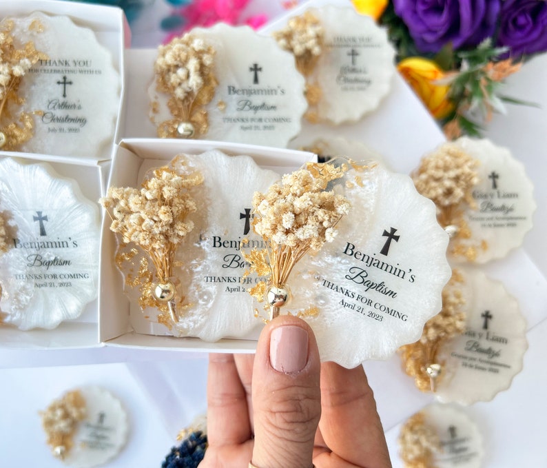 May include: White and gold resin coasters with dried floral accents. Each coaster features a cross and the text "Benjamin's Baptism" and "Thanks for coming April 21, 2023". Some coasters are in a white box.
