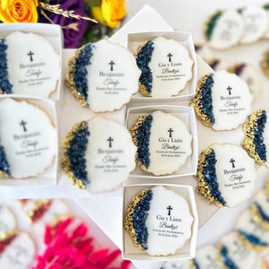 May include: White and gold decorated cookies with blue and gold accents. The cookies have a cross and text that reads "Benjamin Taufe" and "Gia y Liam Bautiz&eacute;".