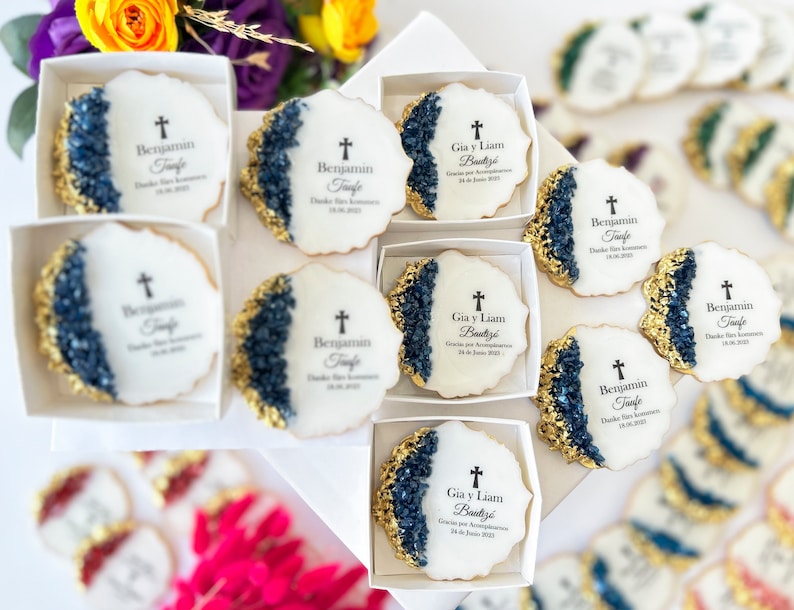 May include: White and gold decorated cookies with blue and gold accents. The cookies have a cross and text that reads "Benjamin Taufe" and "Gia y Liam Bautiz&eacute;".