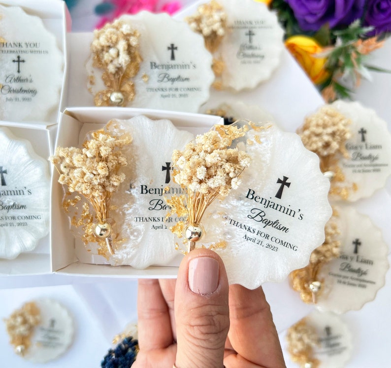 May include: White and gold resin coasters with dried floral accents. Each coaster features a cross and the text "Benjamin's Baptism" and "Thanks for Coming" with a date. Some coasters are in a white box.