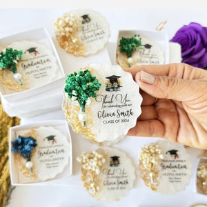 K&ouml;nnte beinhalten: Abschlussgeschenke mit einem Untersetzer aus wei&szlig;em und goldenem Harz mit einer Abschlusskappe und dem Text "Thank You for celebrating with me! Olivia Smith CLASS OF 2024." Jeder Untersetzer ist mit Trockenblumen verziert.