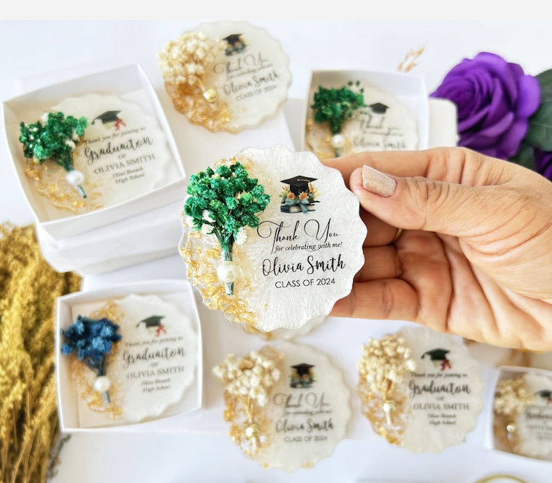 K&ouml;nnte beinhalten: Abschlussgeschenke mit einem Untersetzer aus wei&szlig;em und goldenem Harz mit einer Abschlusskappe und dem Text "Thank You for celebrating with me! Olivia Smith CLASS OF 2024." Jeder Untersetzer ist mit Trockenblumen verziert.