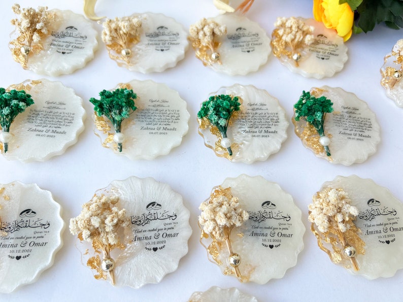 May include: White and gold resin coasters with floral accents. Each coaster features a unique design with gold leaf, dried flowers, and printed text. Some coasters have green floral arrangements, while others have beige. The text includes names and dates.