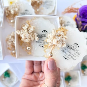 May include: White resin coasters with gold leaf and dried flower decorations. Each coaster has Arabic calligraphy and the names "Amina & Omar" with a date. The coasters are presented in a white box.