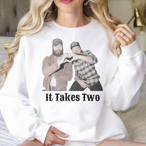 May include: White sweatshirt with a graphic of two men making a heart shape with their hands. The text "It Takes Two" is printed below the graphic.